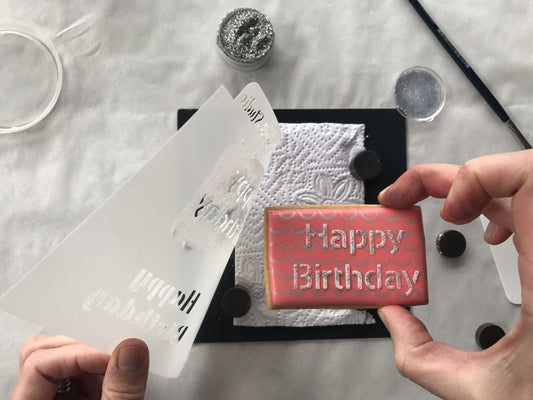 Decorating a rectangular Happy Birthday cookie with stencil, pink icing, and silver glitter