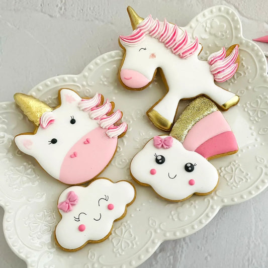 Decorated sugar cookies shaped like unicorns, clouds, and rainbows with pink and gold icing.